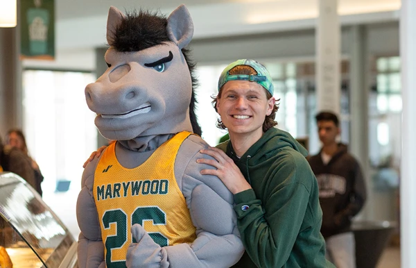 A young man with curly blonde hair wearing a green cap and shirt hugs the Marywood University mascot, a gray horse in a basketball jersey, inside a campus building.